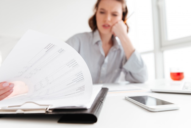 Femme assise à son bureau dans un appartement lumineux, lisant attentivement des documents papier